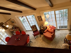 A cozy living room with a high ceiling, exposed beams, a dining area with a red checkered tablecloth, and large windows offering a view of the outside.