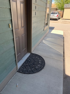 A house entrance with a brown door, a doormat, and a driveway leading to a parked car. - An Apartment Gem Located In The Heart Of Missoula (Missoula)