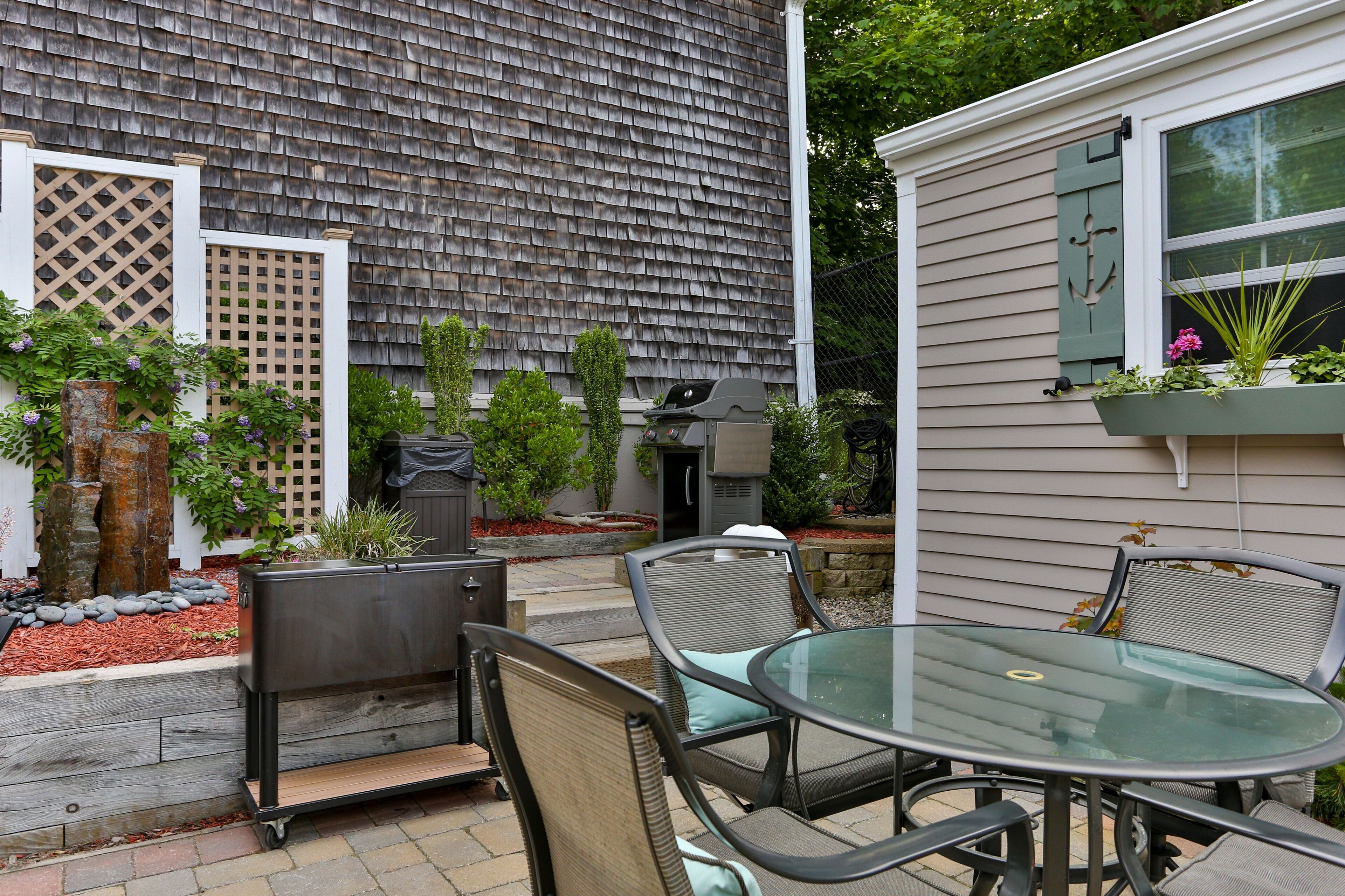 A patio with a glass table and chairs, a barbecue grill, and a small garden area with plants and a wooden fence.
