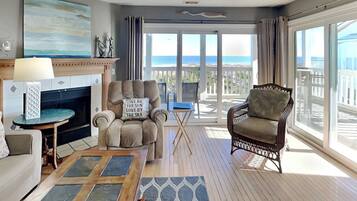 A spacious living room with a fireplace, a comfortable armchair, a sofa, and a view of the ocean through large windows.
