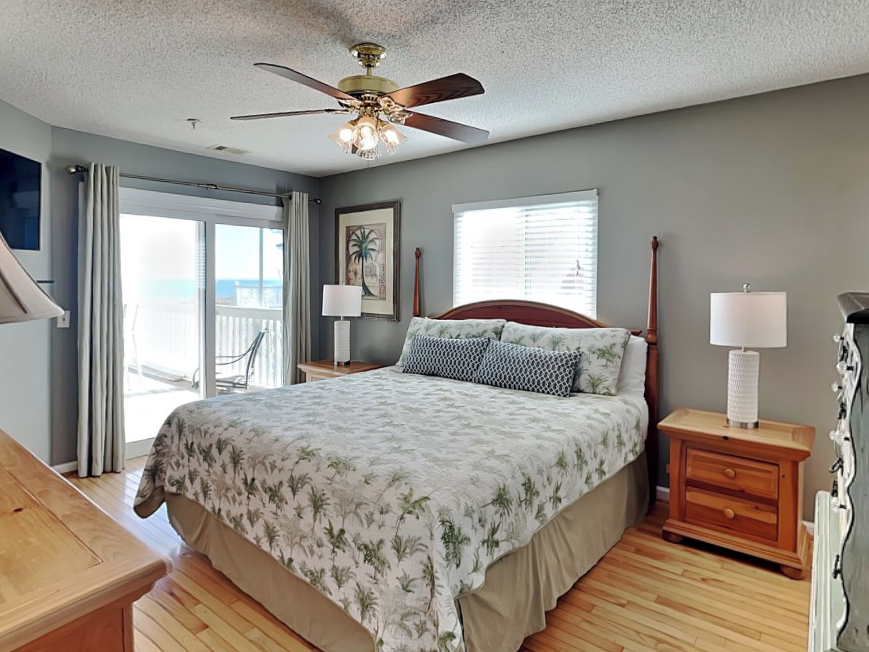 A bedroom with a bed, a ceiling fan, a nightstand with a lamp, a wooden dresser, and a view of the outdoors through a sliding door.