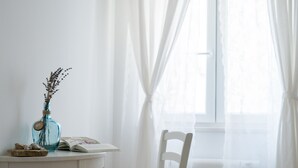 A room with a white chair, a small table with a vase and a book, and a window with sheer curtains.
