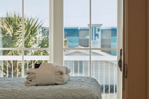A neatly made bed with a folded towel on it, a balcony with a railing, and a view of buildings and palm trees outside.