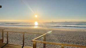 A beach with a wooden railing, waves, and the sun setting or rising over the ocean.