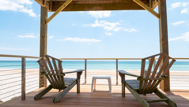 A wooden deck with two rocking chairs and a small stool, overlooking a beach with clear blue water and a wooden railing.
