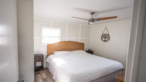 A bedroom with a large bed, a ceiling fan, a bedside table, a nightstand, and a wall-mounted decoration.