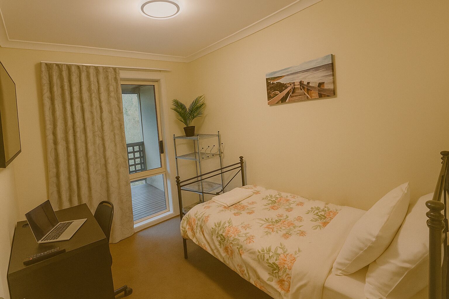 A bedroom with a bed, a desk with a laptop, a window with curtains, a plant on a metal shelf, and a painting on the wall.
