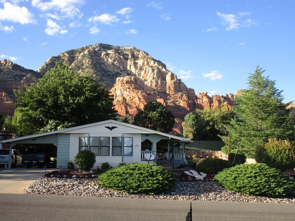 A small building with a mountain backdrop, surrounded by greenery and a gravel driveway.
