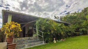 Una casa de piedra con un porche cubierto y un jardín exuberante con diversas plantas y una maceta.