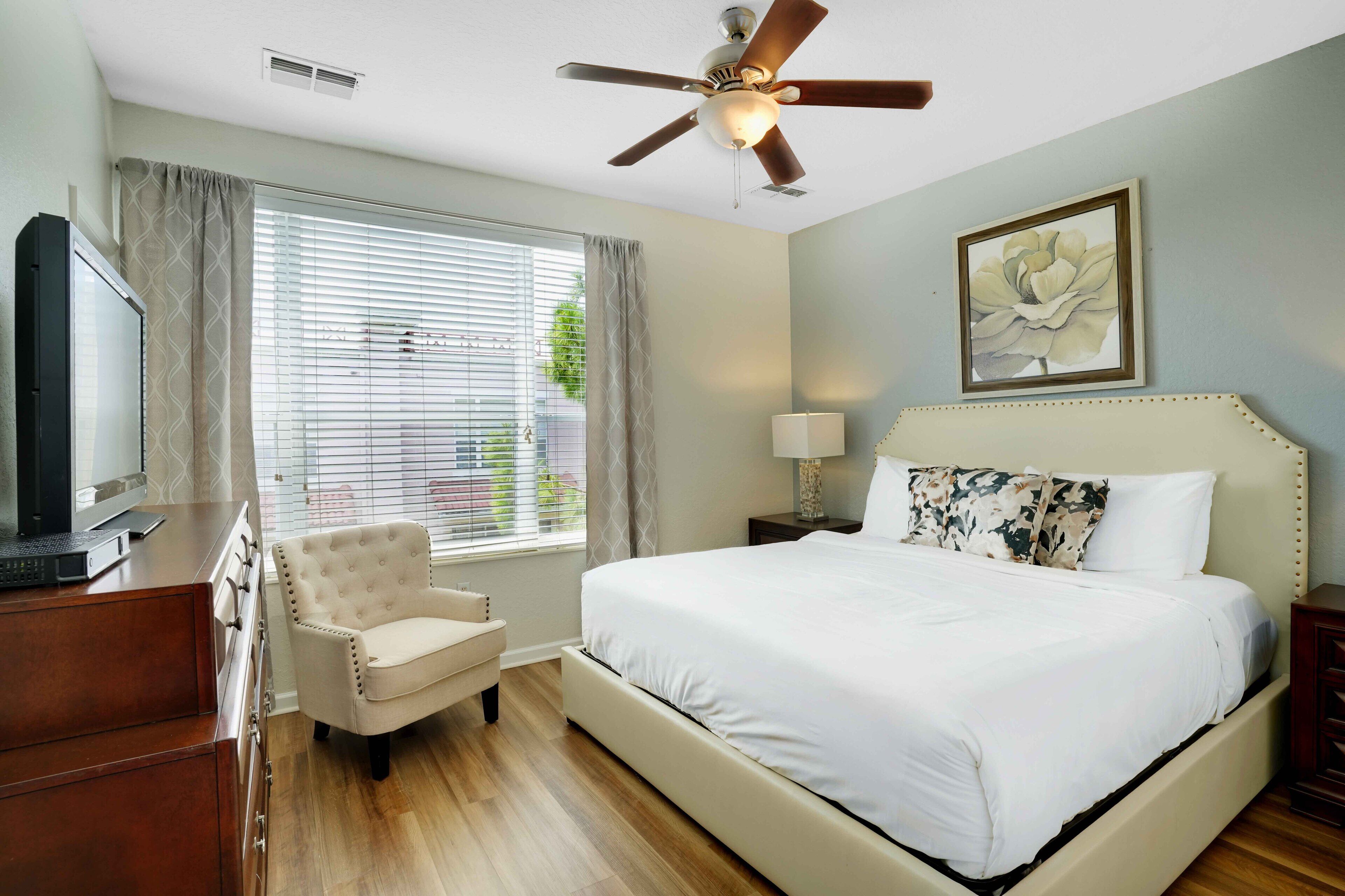 A hotel room with a bed, a chair, a desk with a television, a ceiling fan, and a window with curtains.