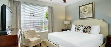 A hotel room with a bed, a chair, a desk with a television, a ceiling fan, and a window with curtains.