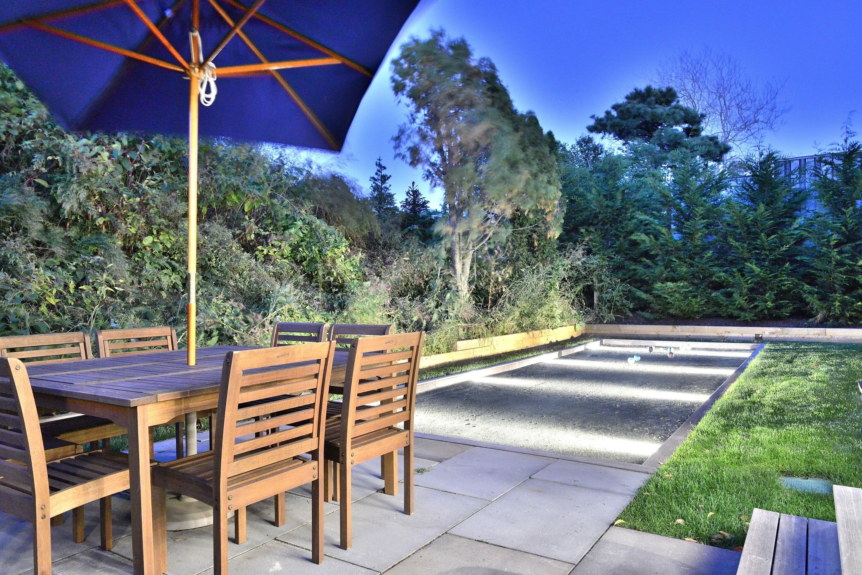 An outdoor patio with a wooden table and chairs, a blue umbrella, and a well-maintained lawn.
