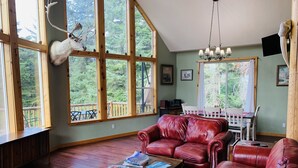 Ein geräumiges Wohnzimmer mit einem roten Ledersofa, einem Couchtisch aus Holz mit Büchern und einem großen Fenster, an dessen Wand ein Hirschkopf angebracht ist.