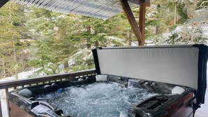 Ein Whirlpool mit Holzverkleidung und Metallabdeckung, umgeben von schneebedeckten Bäumen.