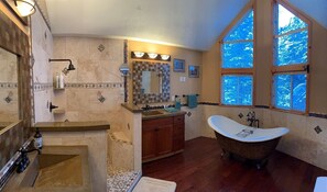 Et badeværelse med et fritstående badekar, et toiletbord med spejl og et vindue med udsigt til træer.