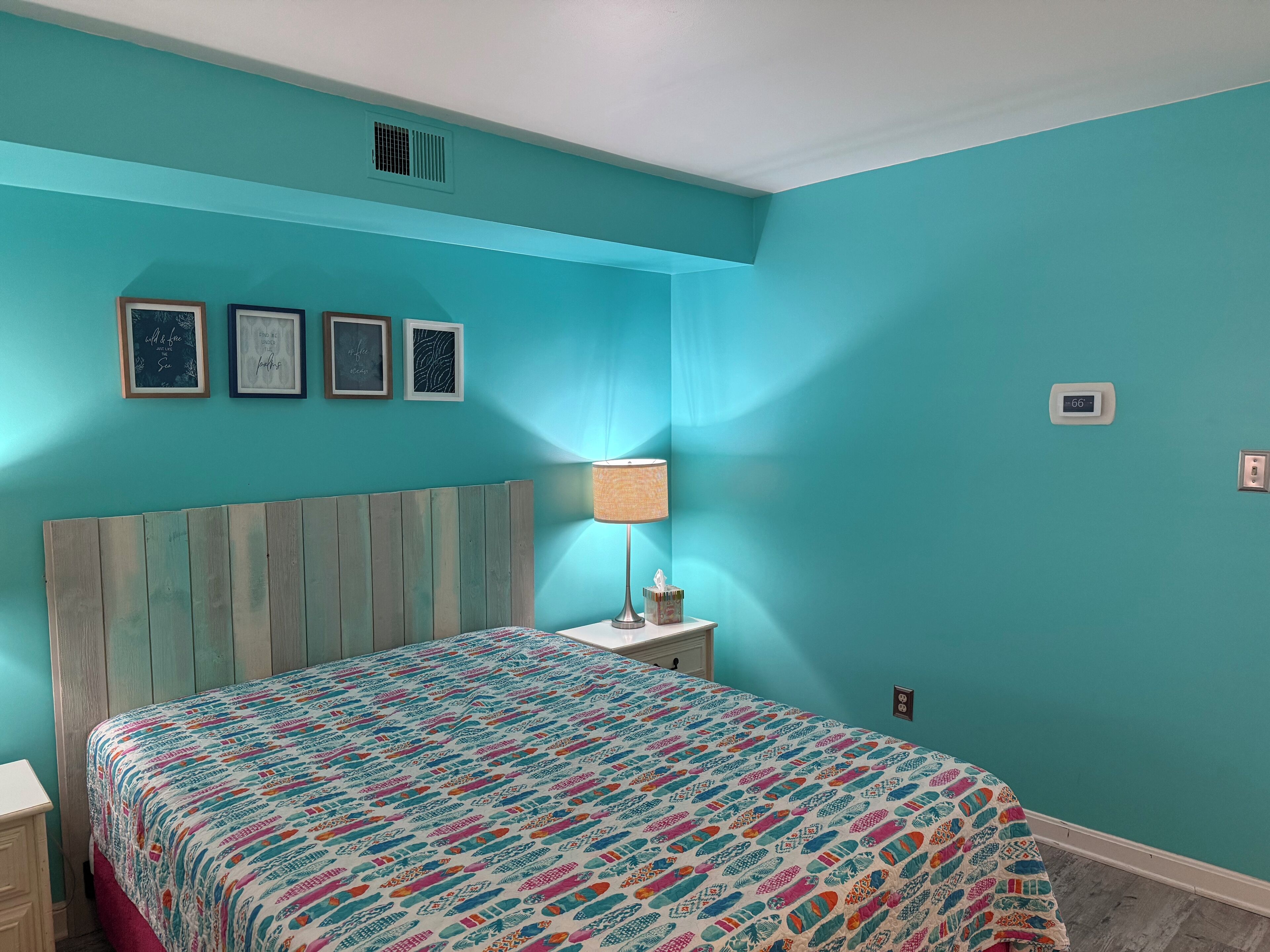 Un dormitorio con paredes color turquesa, cabecera de madera, colcha colorida, una lámpara de noche y tres cuadros en la pared.
