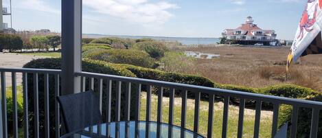 A glass table and chair on a balcony with a view of a beach and a building.