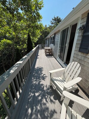 Una terraza de madera con sillas Adirondack, una mesa y un ventanal que da a un área cubierta.
