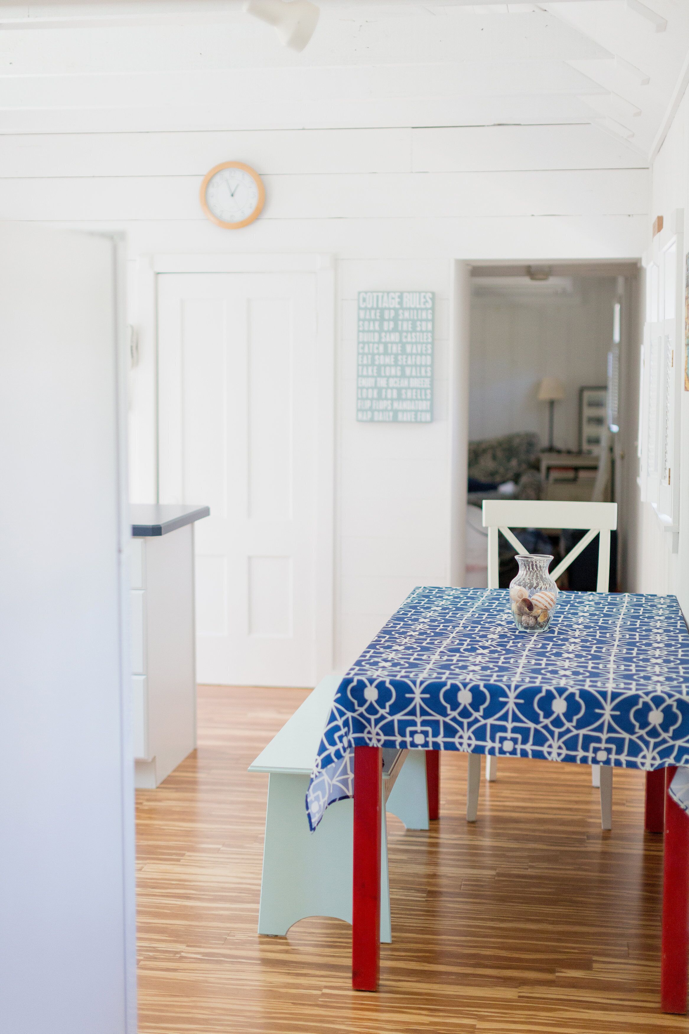 Une pièce avec un plancher en bois, une porte blanche, une horloge au mur et une table recouverte d’une nappe à motifs.