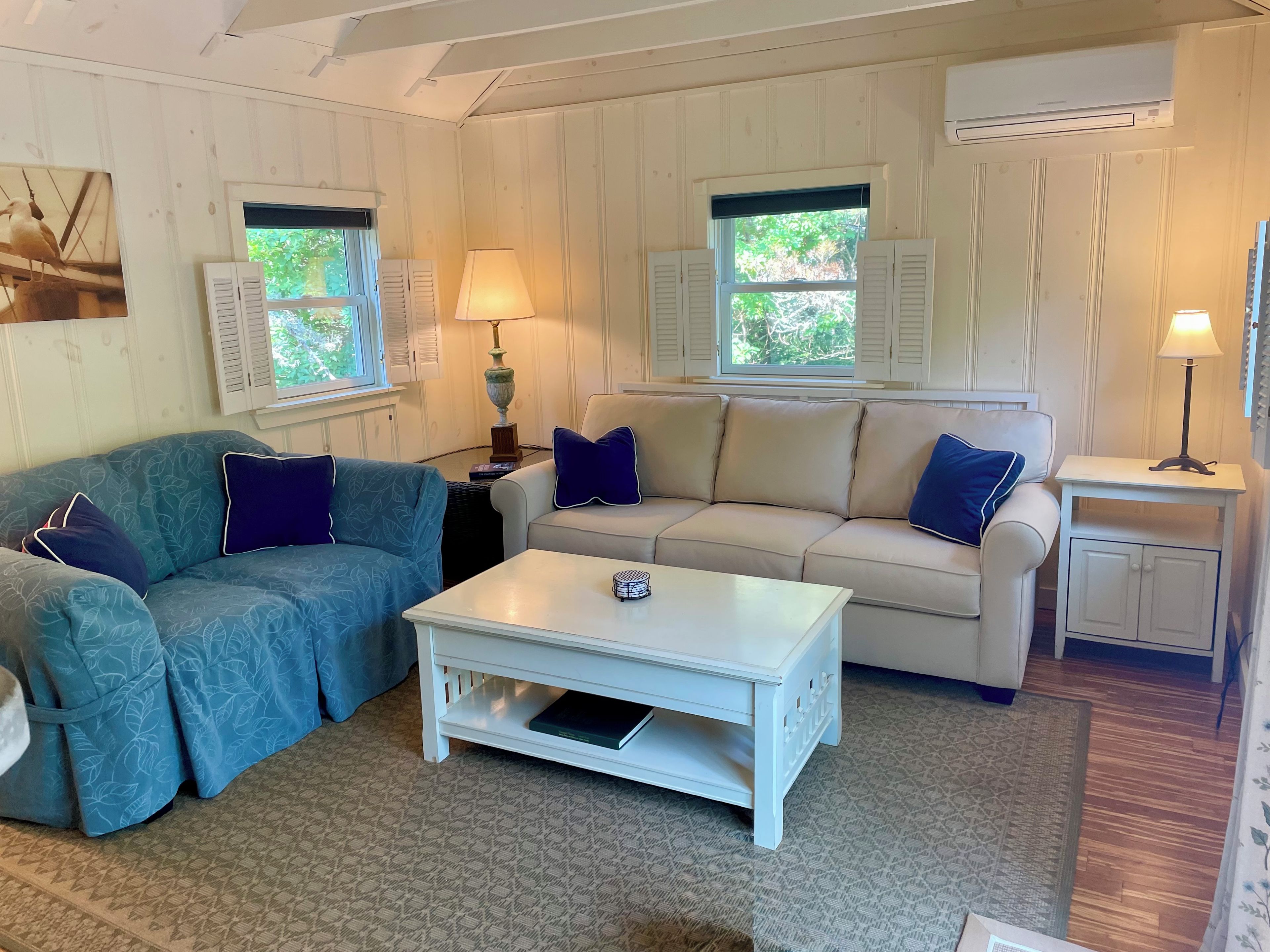 A cozy living room with a blue patterned sofa, a white coffee table, and a beige sofa with blue pillows. There are two windows with shutters, a lamp on a side table, and an air conditioning unit on the wall.