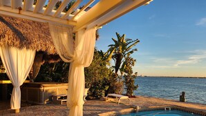 Un espace piscine offrant une vue dégagée sur l’eau, une pergola agrémentée de rideaux blancs et un coin salon doté d’un toit de chaume.