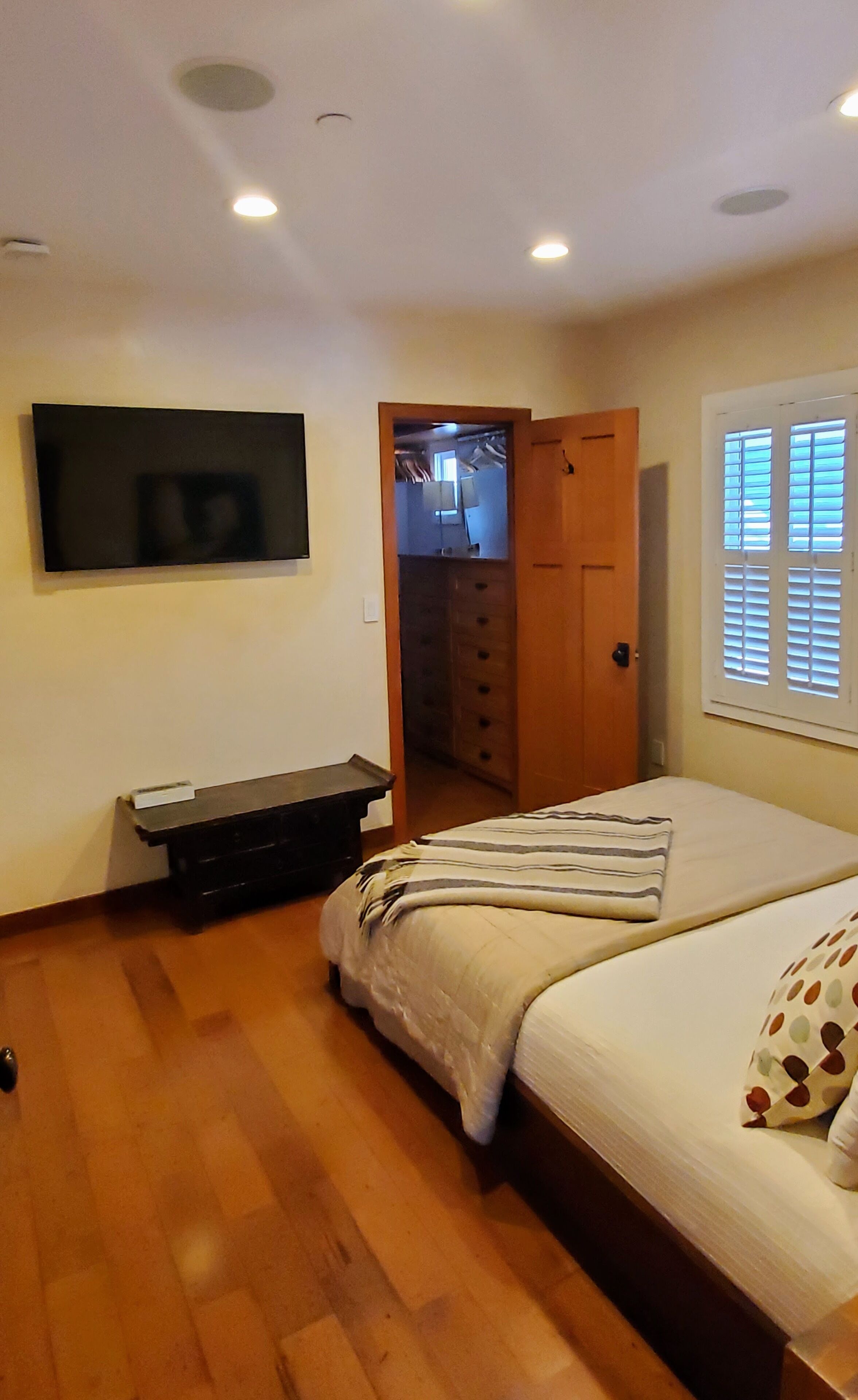 A bedroom with a bed, a TV mounted on the wall, a wooden dresser, and a closet with a door.