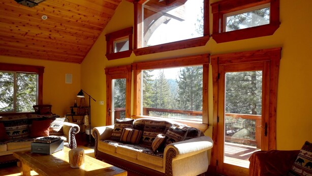 A cozy cabin interior with a wooden ceiling, large windows, a sofa with patterned cushions, a wooden table, and a chair. - Tahoe Ski Lodge  WiFi, hot-tub sleeps 12 (Truckee)