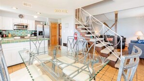 A modern kitchen with a central island, a staircase with a cat tree, and a living area with a sofa and lamp. - On Beach, Sunsets, Bikes+Kayaks, Pet Friendly, Firepit, Serenity.  (Villas)