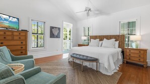 A bedroom with a large bed, a television, a ceiling fan, and a view of the outdoors through the windows.