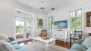 A spacious living room with a white ceiling fan, a large flat-screen TV, and a view of the outdoors through large windows.