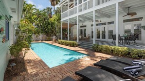 A pool area with lounge chairs, a covered patio, and a view of a building with a balcony.