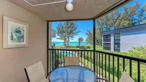A balcony with a glass table and chairs, a framed painting on the wall, and a view of the ocean and a building across the street. - Sea Grape Inn #5- Beachfront Property (Longboat Key)