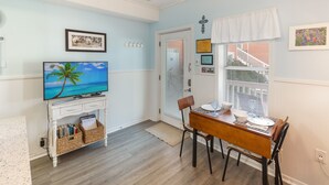 A dining area with a wooden table and chairs, a television on a white console, and a window with blinds. - #110 Picture Perfect Studio w Sleeper Sofa & Pool (Isle of Palms)