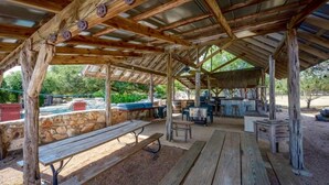 An outdoor covered patio area with wooden benches and tables, stone walls, and a barbecue setup. - 16-Acre Resort Ranch with 4 Homes Included, Pool, Spa, Firepit &Gathering Spaces (Kyle)