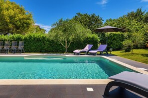 Une piscine à l’eau claire, entourée d’une terrasse dallée. On y trouve des chaises longues et un parasol.