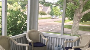 A covered porch with wicker chairs and a drum table, overlooking a street with houses and trees. - Adorable Beach Bungalow - Awesome location! Walk to beach and town! (South Haven)