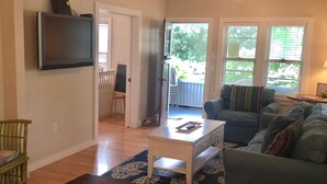 A living room with a television, a coffee table, a sofa, and a dining table with a plate of tomatoes. - Adorable Beach Bungalow - Awesome location! Walk to beach and town! (South Haven)