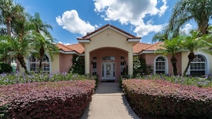 A two-story building with a red-tiled roof, a central entrance with a glass door, and symmetrical landscaping with hedges and palm trees.