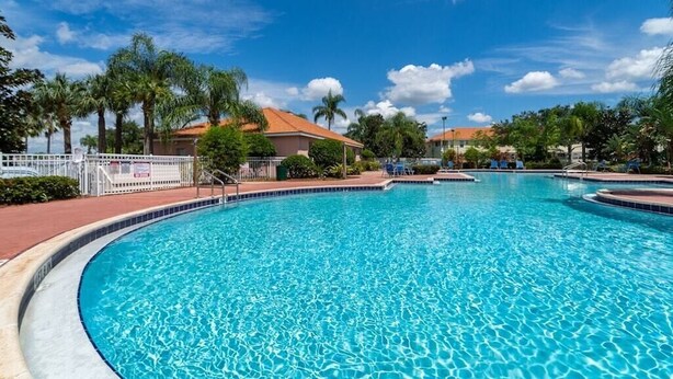 A large, clear swimming pool with a curved edge, surrounded by palm trees and a building with a red roof.