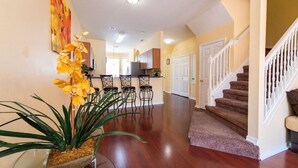 A spacious living area with a wooden floor, a dining area with bar stools, a staircase, and a kitchen in the background.