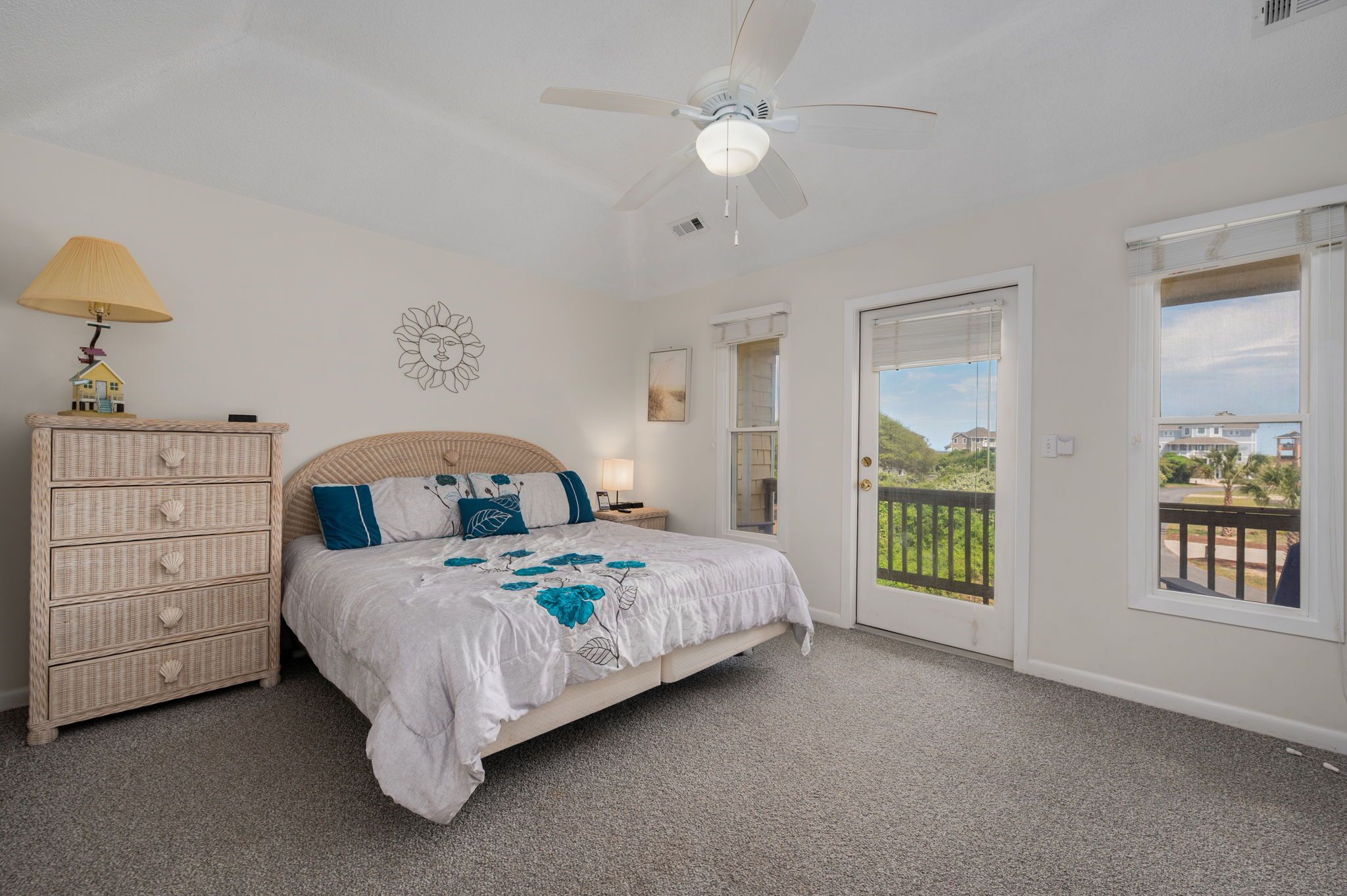 A bedroom with a bed, a ceiling fan, a dresser, a nightstand, a lamp, a window, and a balcony.