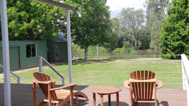 Una terraza con dos sillas de madera tipo Adirondack y una pequeña mesa, con vista a un área verde con agua y árboles.