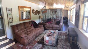 A cozy living room with a brown leather sofa, a matching armchair, and a coffee table with a patchwork cover. A wooden ladder decorates the wall, and a 'Welcome' sign hangs on a corrugated metal door.