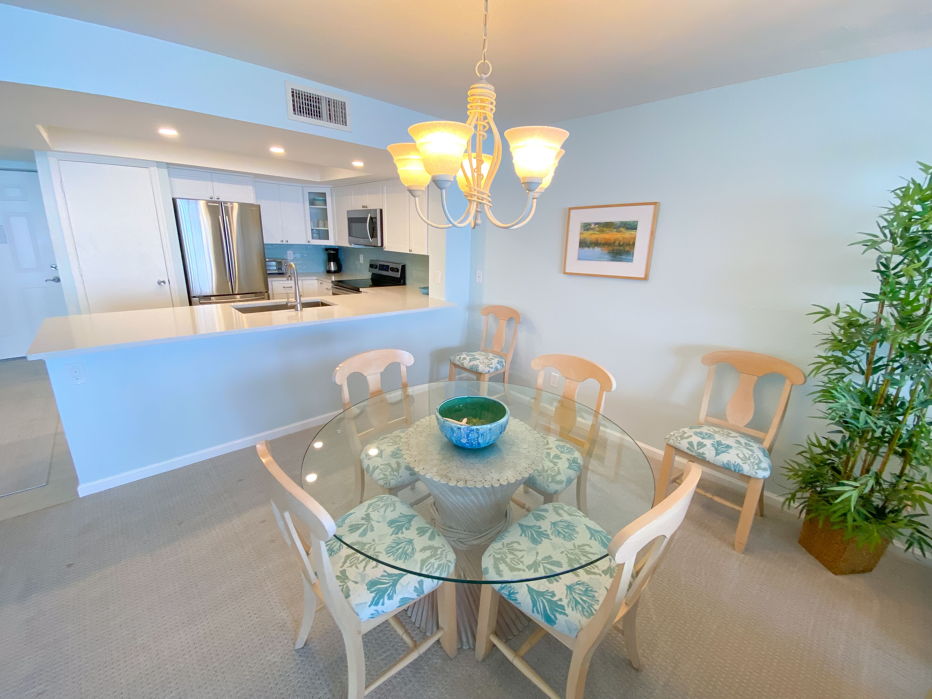 A modern kitchen with a central island, a dining area with a glass table and chairs, and a chandelier.