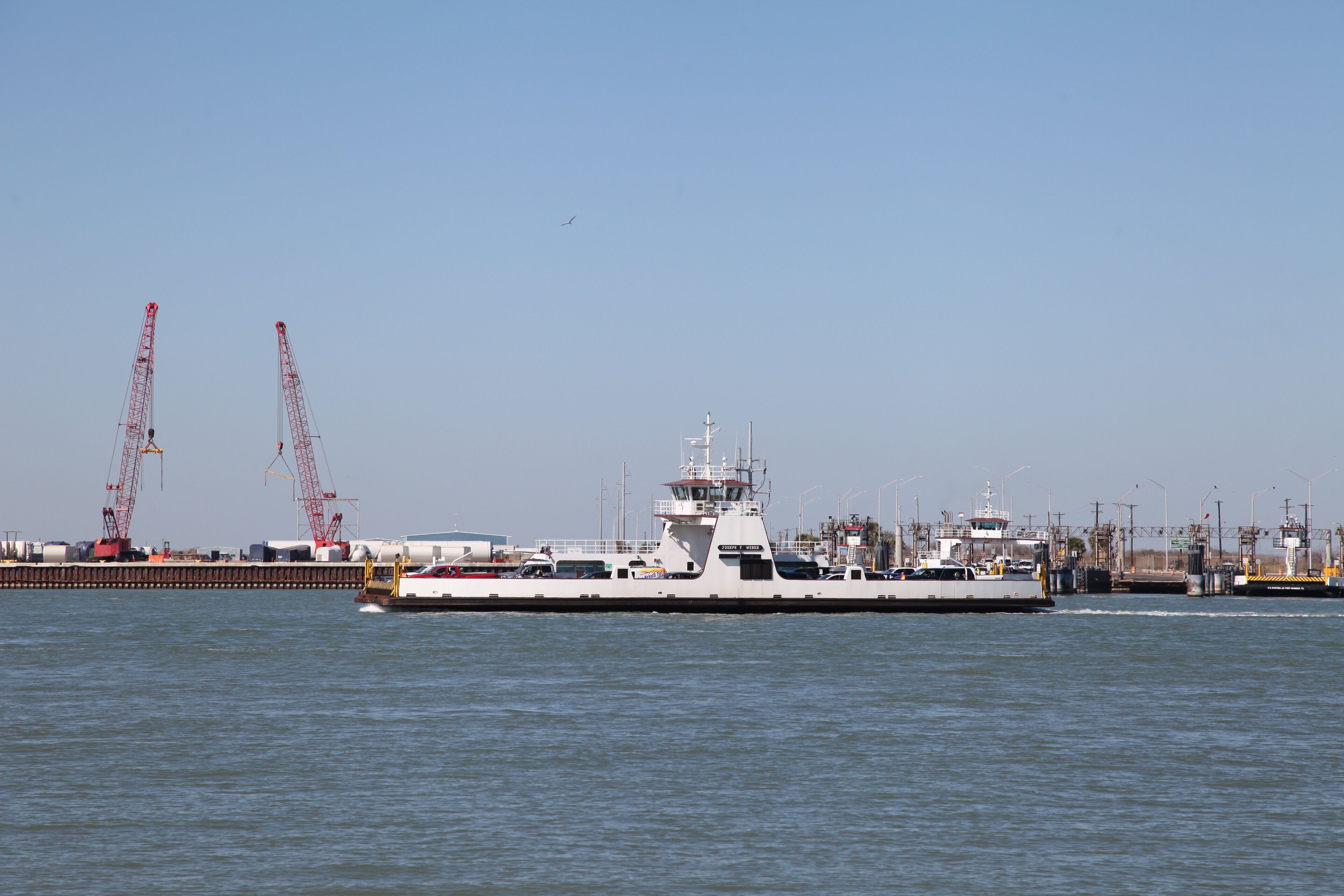 A large white ship docked at a port with cranes and industrial structures in the background.