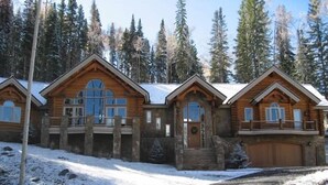 A log cabin with a large porch and a garage, surrounded by a snowy landscape and tall trees. - 5 BR + Loft Luxury Ski-in & Ski-out Log Home, stunning mountain views, hot tub (Mt Village)