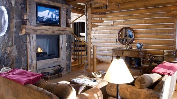 A log cabin interior with a stone fireplace, a television mounted above it, and a cozy seating area with a lamp. - 5 BR + Loft Luxury Ski-in & Ski-out Log Home, stunning mountain views, hot tub (Mt Village)