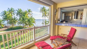 A balcony with two wooden lounge chairs and a small table, overlooking a beach with palm trees and the ocean.