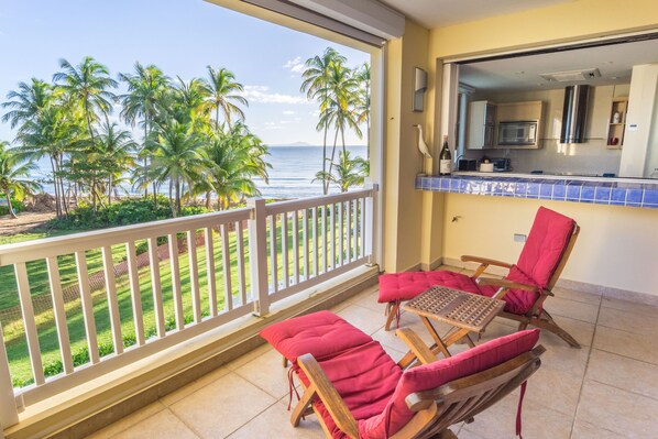 A balcony with two wooden lounge chairs and a small table, overlooking a beach with palm trees and the ocean.