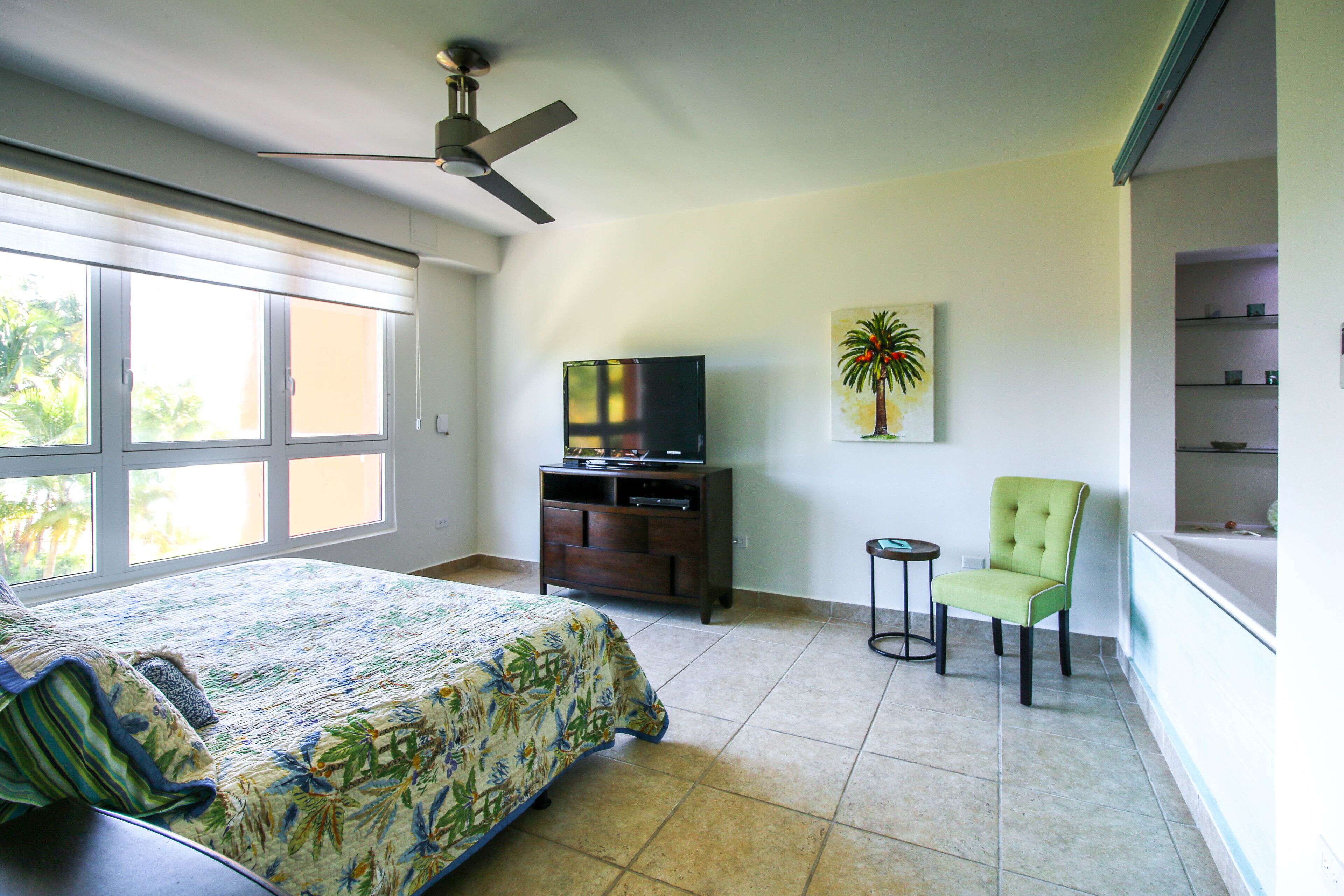 Un dormitorio con cama, televisión sobre un mueble, una silla y un cuadro con una palmera.